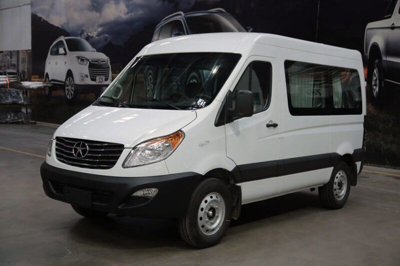 Белый пассажирский микроавтобус JAC Sunray, вид спереди и сбоку, стоит в выставочном зале на фоне других автомобилей JAC.