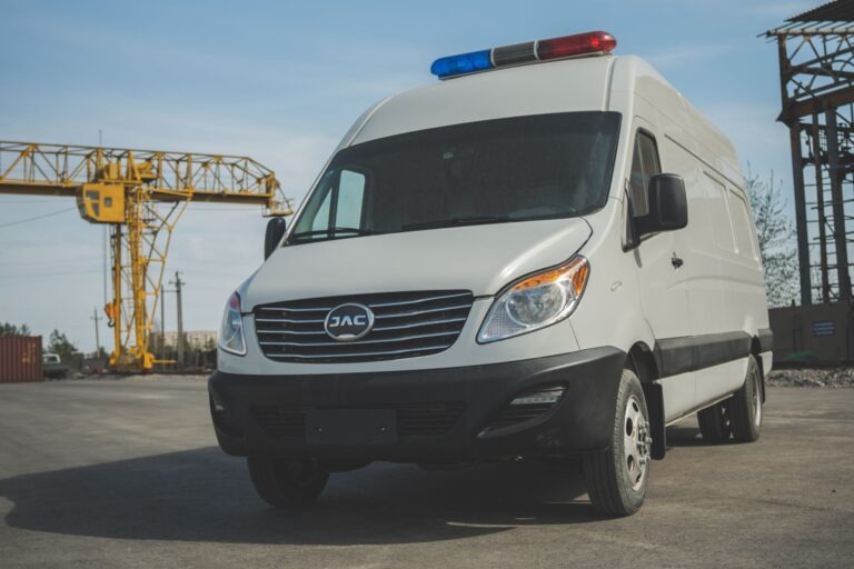 Белый фургон JAC Sunray с мигалкой на крыше, вид спереди, стоит на промышленной площадке на фоне крановых конструкций.